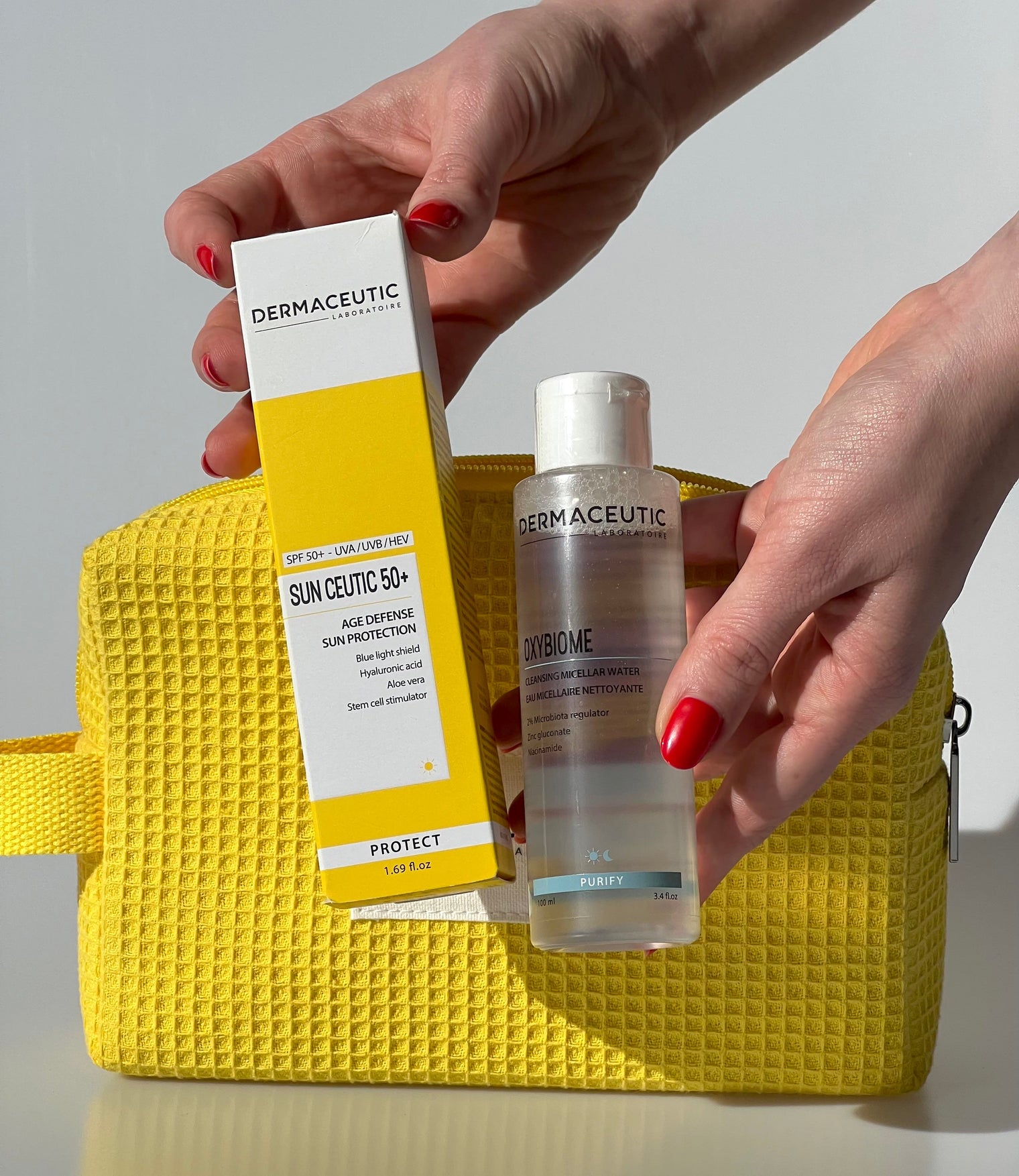 Two skincare products from Dermaceutic held by hands against a yellow textured bag.